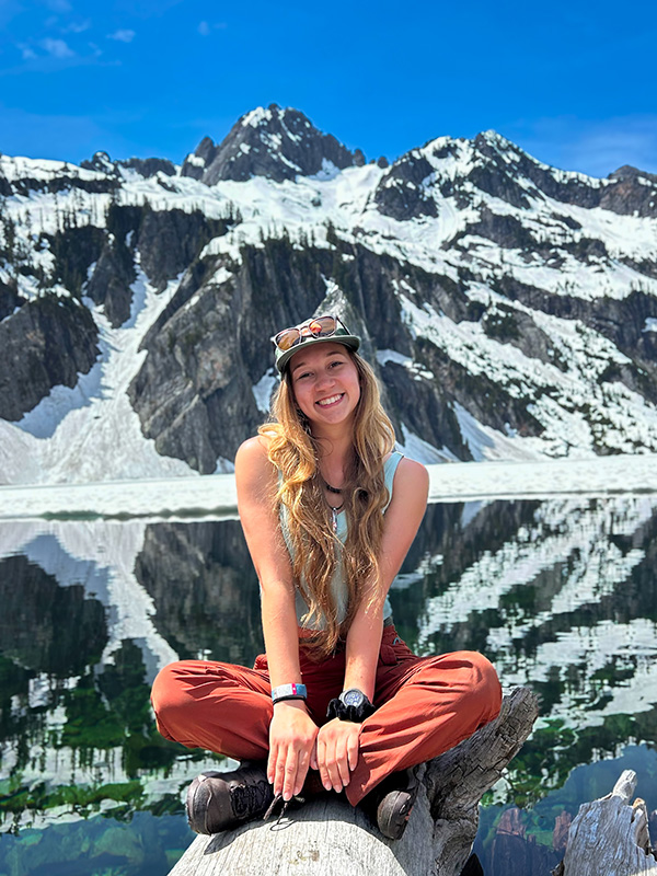 Photograph of a young woman sitting cross legged on a log with a mountain backdrop