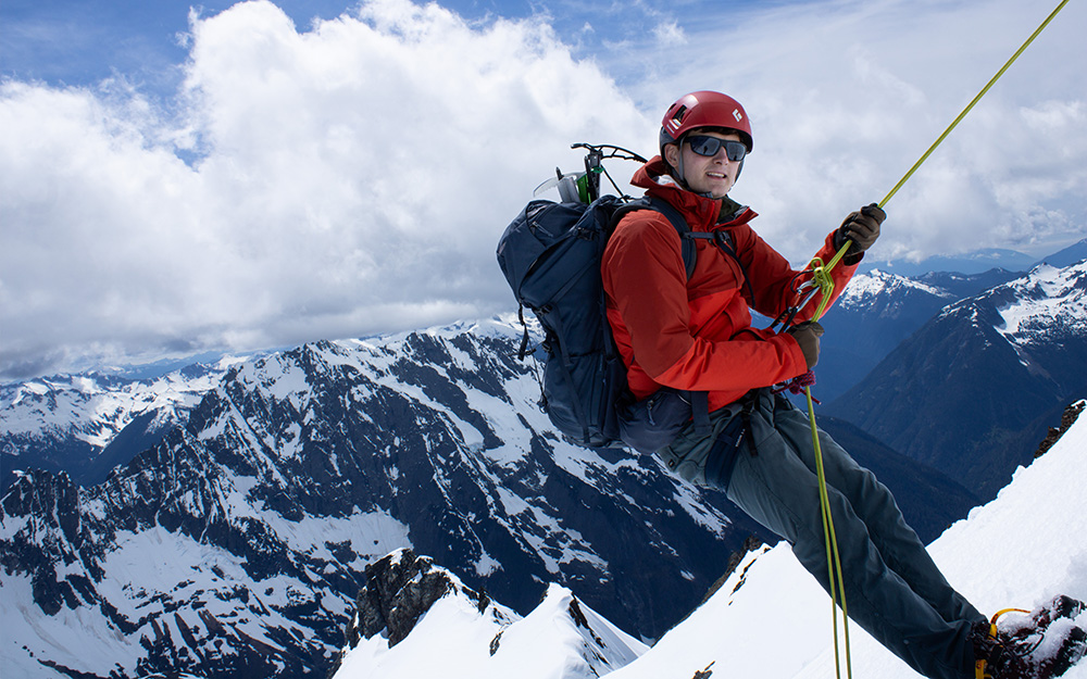 Male mountaineer in red rappeling off a summit block with the North Cascades in the background