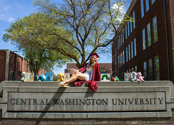 A girl in a red cap and gown sits with stuffed animals of childrens book characters on her college sign