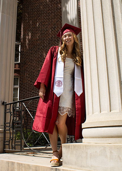 A girl in a white dress and a red cap and gown leans against a white pillar
