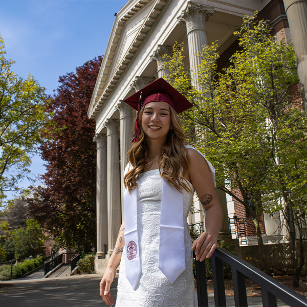 A girl in a white dress and graduation cap stands by a rail in front of a college building