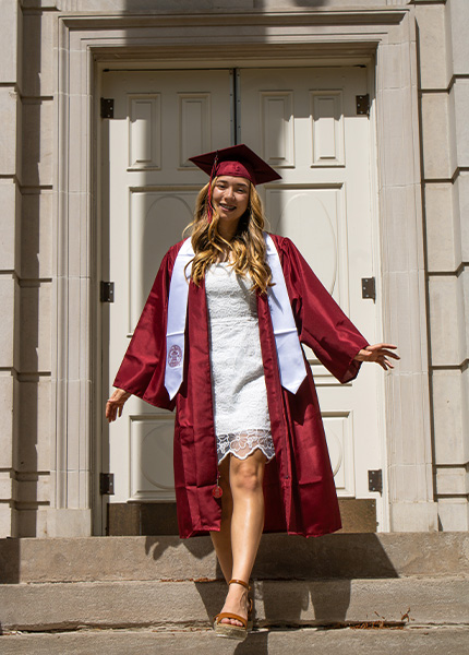 A girl in a white dress and a red cap and gown steps down a set of stairs