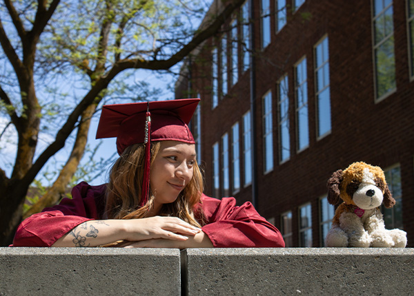 A girl in a white dress and a red cap and gown looks at her childhood stuffed animal next to her