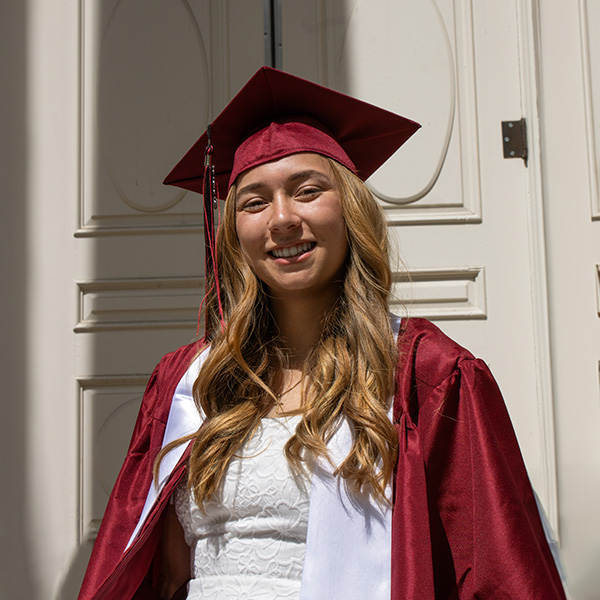 A girl in a white dress and red cap and gown smiles for a photo
