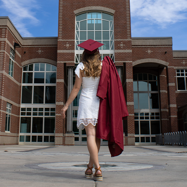 A girl in a white dress and a red cap and gown walks towards a building