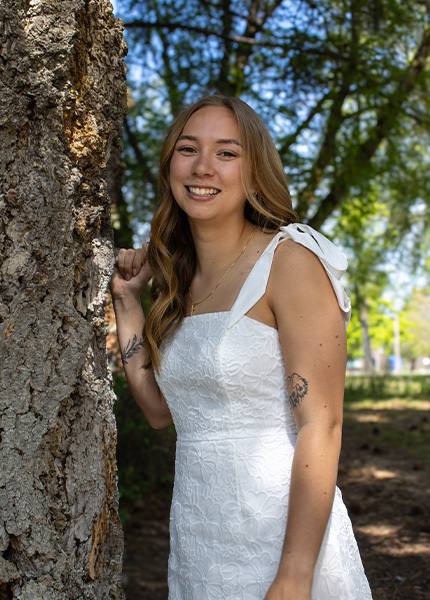 A smiling girl in a white dress leans against a tree