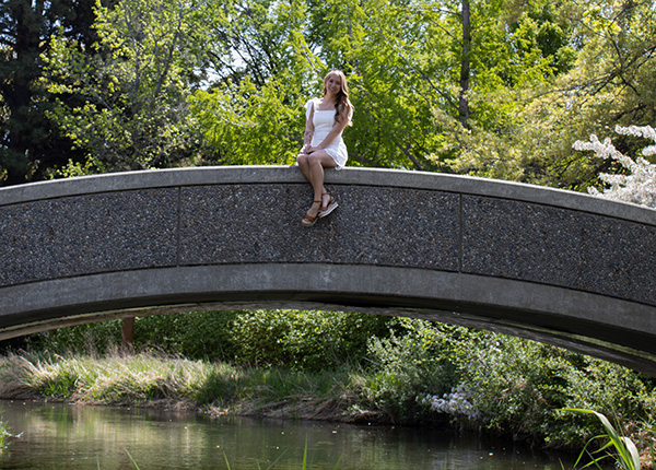 A girl in a white dress sits on a small concrete bridge over a river