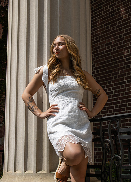 A girl in a white dress with blonde hair leans against a pillar