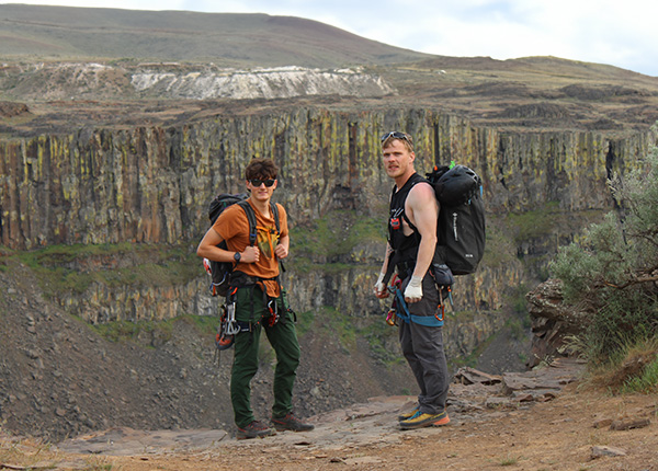Two men stand in front of a cliff with backpacks and rock climbing gear