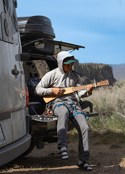 A young man sits on the tailgate of a truck playing a guitar with a climbing harness on